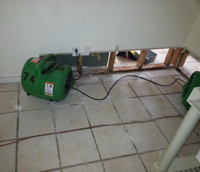 Two green air movers drying a wall cavity on tile flooring.  The wall has a flood cut in it.