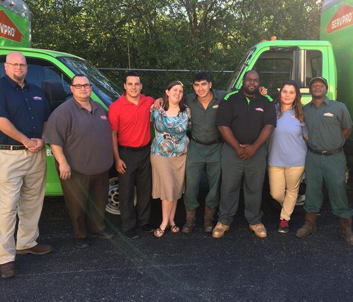 Six men and two women standing together smiling in front of two green box trucks