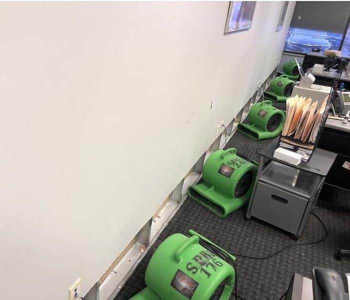 An office with drywall removed on the lower part of a wall, with SERVPRO equipment on the carpeted flooring. 
